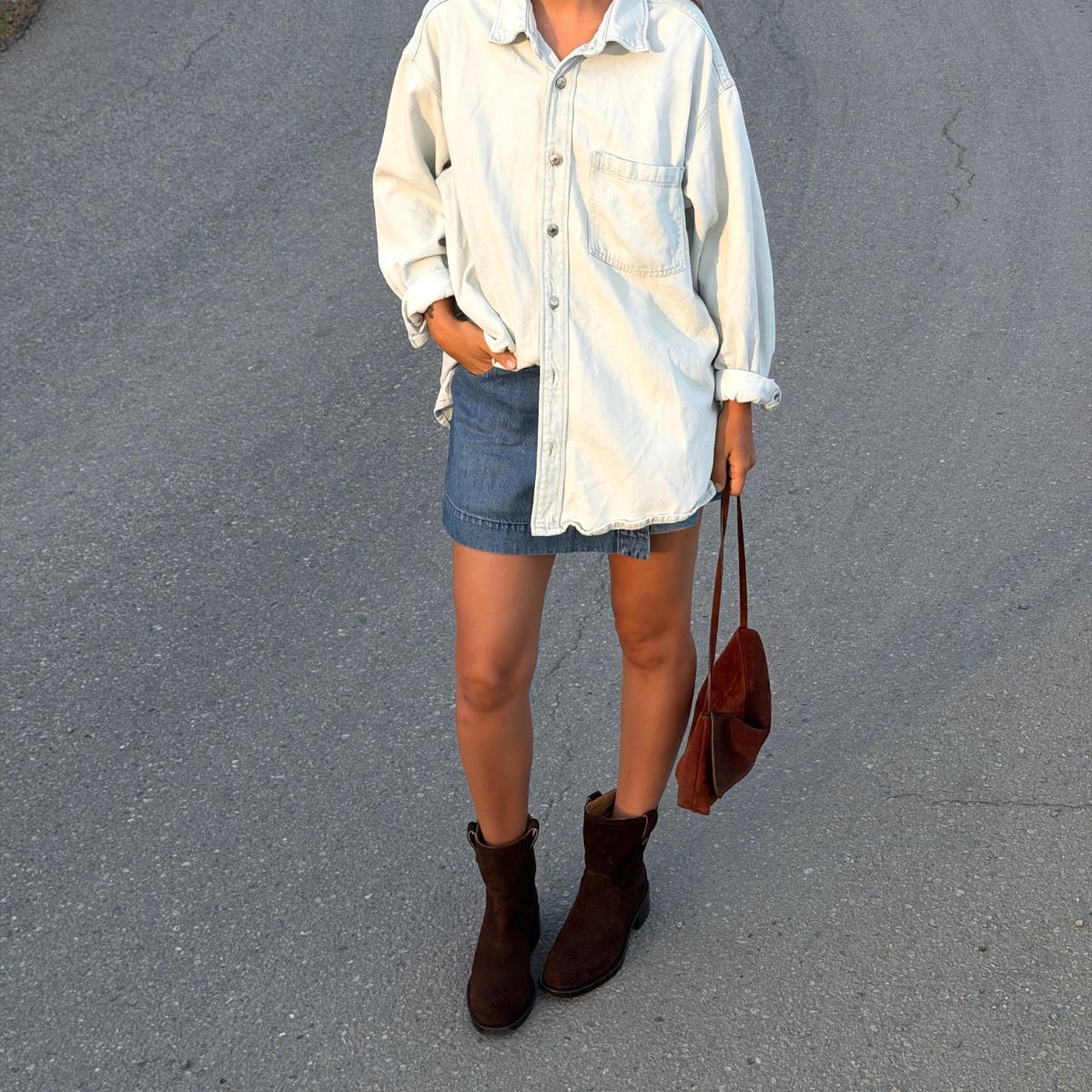 A person wearing brown suede Viola Moto boots, a denim skirt, and a light blue button-down shirt standing on asphalt.